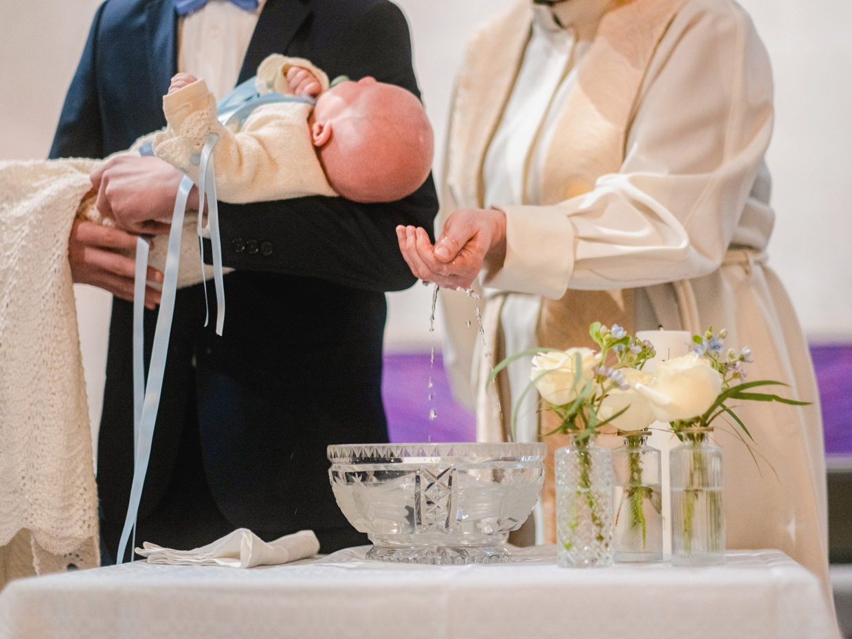 Mieshahmo pitelee sylissään kastemekkoon puettua vauvaa papin nostaessa vettä kastemaljasta.