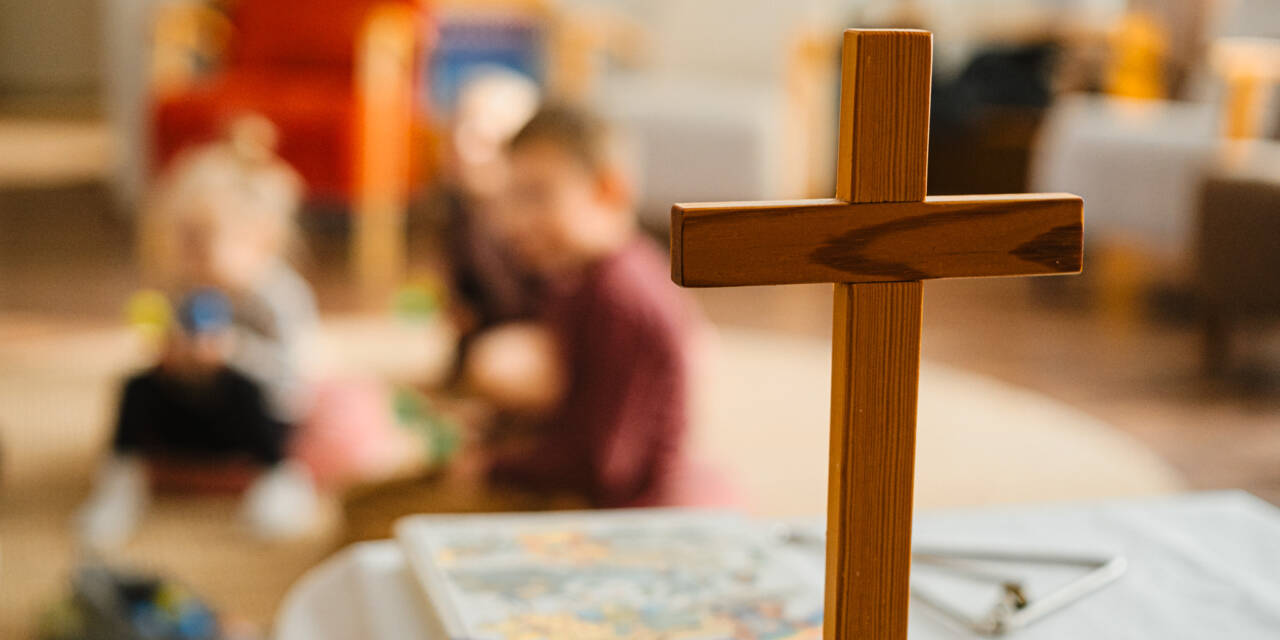 Pöydälle pystytetty puinen risti, jonka taustalla näkyy sumeana lattialla istuvia lapsia.