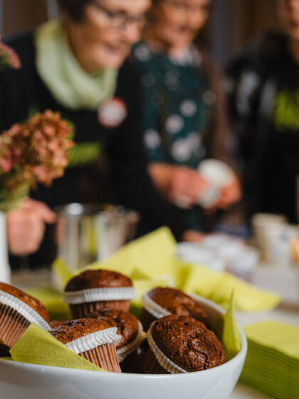 Etualalla kulho, jossa on muffineja. Taustalla näkyy sumeasti kaksi rinnakkain pöydän ääreen kumartunutta naishahmoa. Pöydällä on astioita.