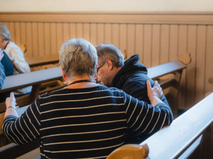 Kaksi takaapäin kuvattua ihmistä istuu vieretysten kirkonpenkissä aavistuksen eteenpäin ja toisiaan kohti kumartuneina. Toinen pitää kättään kevyesti toisen selällä.