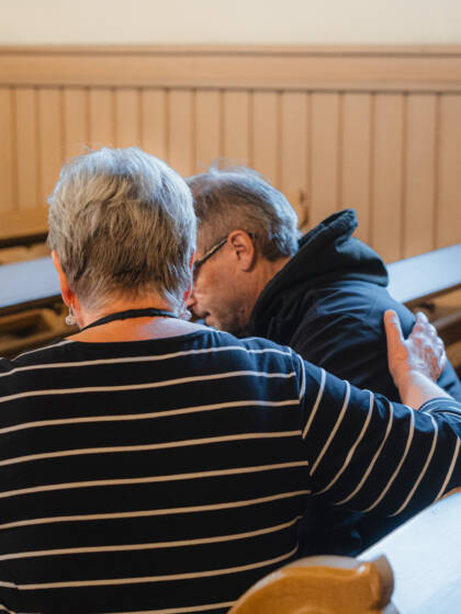 Kaksi takaapäin kuvattua ihmistä istuu vieretysten kirkonpenkissä aavistuksen eteenpäin ja toisiaan kohti kumartuneina. Toinen pitää kättään kevyesti toisen selällä.