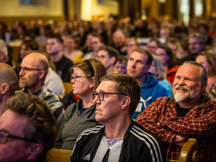 Edestäpäivn kuvattuja ihmisiä istumassa kirkossa. Sali vaikuttaa täydeltä.