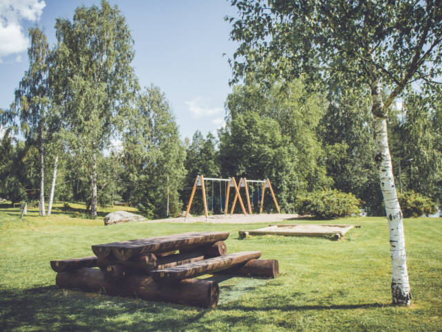 Pyysalon leirikeskuksen pihaa kesällä. Edustalla näkyy pöytä pitkine penkkeineen, taempana keinut.