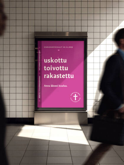 Jonkin julkisen tilan käytävällä sähköinen ilmoitustaulu, jolla näkyy kirkkaana seurakuntavaalien purppurapohjainen mainos ja kampanjansanat: uskottu, toivottu, rakastettu. Ilmoitustaulun ohi kulkee ihmisiä.
