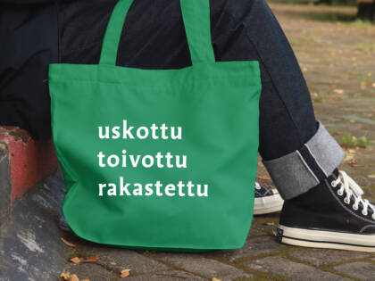 Istuvan ihmishahmon vieressä vihreä kangaskassi, jossa lukee kampanjasanat: uskottu, toivottu, rakastettu.