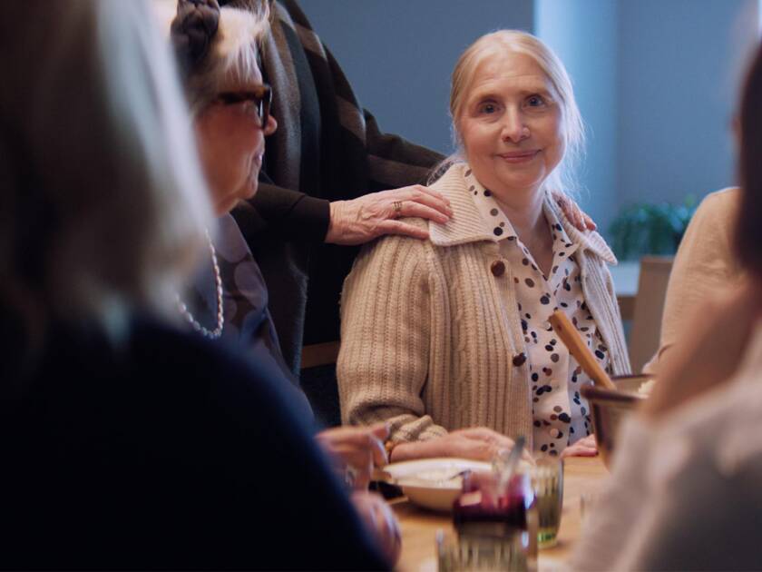 Nainen istuu ruokapöydässä toisten seurassa. Hän hymyilee lempeästi kameraan, ja toinen henkilö laskee rohkaisevasti kätensä hänen olkapäälleen. Tilanne välittää yhteisöllisyyttä ja tukea. Yhteisvastuun kampanjakuva (2026) (c) Yhteisvastuukeräys, Kirkkopalvelut ry