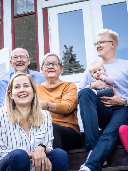 Kuistin rappusilla istuvia eri-ikäisiä ihmisiä vauvasta senioriin. Ihmiset hymyilevät ja katsovat kukin omiin suuntiinsa,