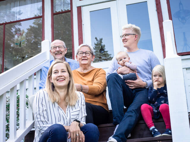 Kuistin rappusilla istuvia eri-ikäisiä ihmisiä vauvasta senioriin. Ihmiset hymyilevät ja katsovat kukin omiin suuntiinsa,