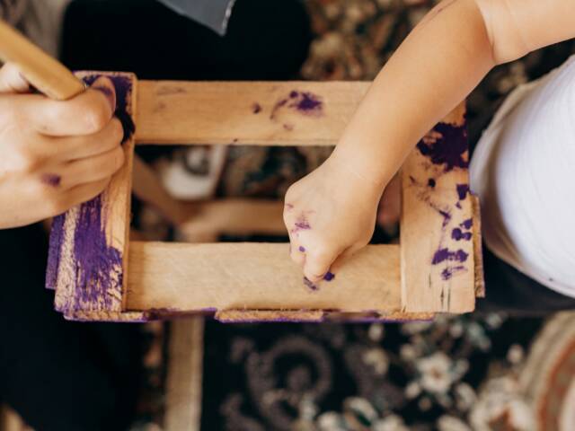 Läheltä otettu valokuva puisesta hyllyköstä tai muusta käsityöstä, jonka ylle kaksi ihmistä on asettunut violettiin maaliin kastettujen pensseleiden kanssa.