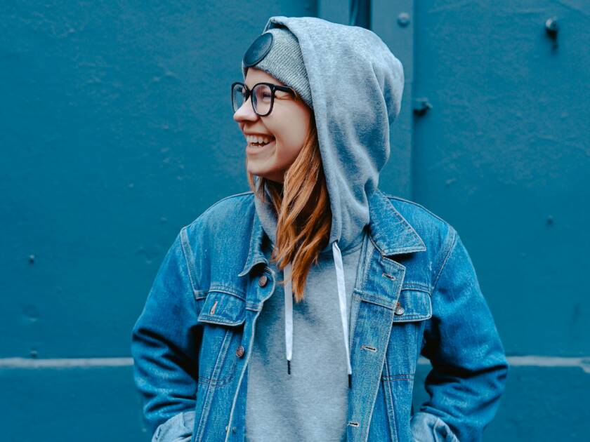 Nuori nainen seisoo kädet farkkutakin taskuissa ja hymyilee sivulle katsoen. Naisella on yllään farkkutakin alla collegehuppari, jonka huppu on vedetty naisen pipon peittämän pään päälle.