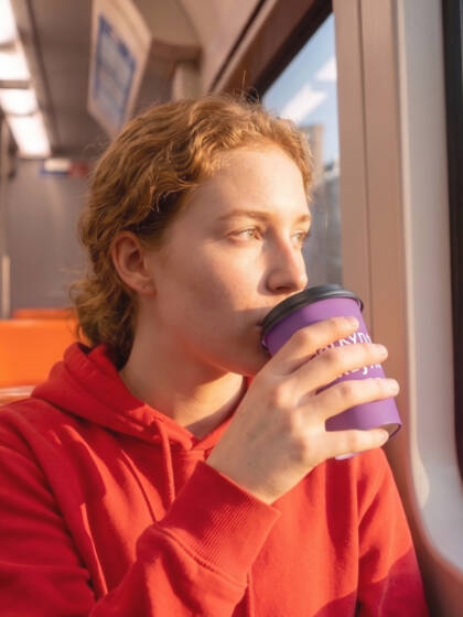 Julkisessa liikennevälineessä istuva nainen siemaisee violetista kahvimukista katsellen samalla ulos ikkunasta.