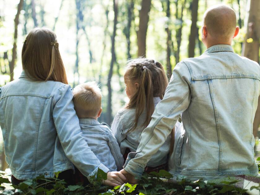 Neljä ihmistä takaapäin kuvattuna: mies ja nainen istuvat maassa kasvot suunnattuna utuisena näkyvää puustoa kohti pitäen toisiaan kädestä. Käsien edessä, aikuisten välissä, istuu kaksi lasta. Kaikilla on yllään samanväriset takit.