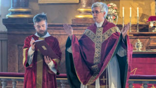 Liturgi tervehtii seurakuntaa punaisessa messukasukassa alidiakoni vierellään.