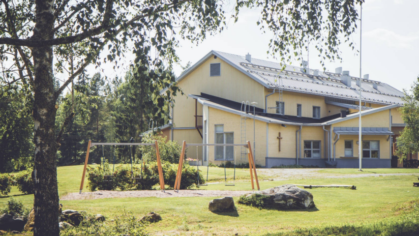 Pyysalo - keltainen iso puutalo iltapäivän auringossa. Etualalla leikkikenttä.