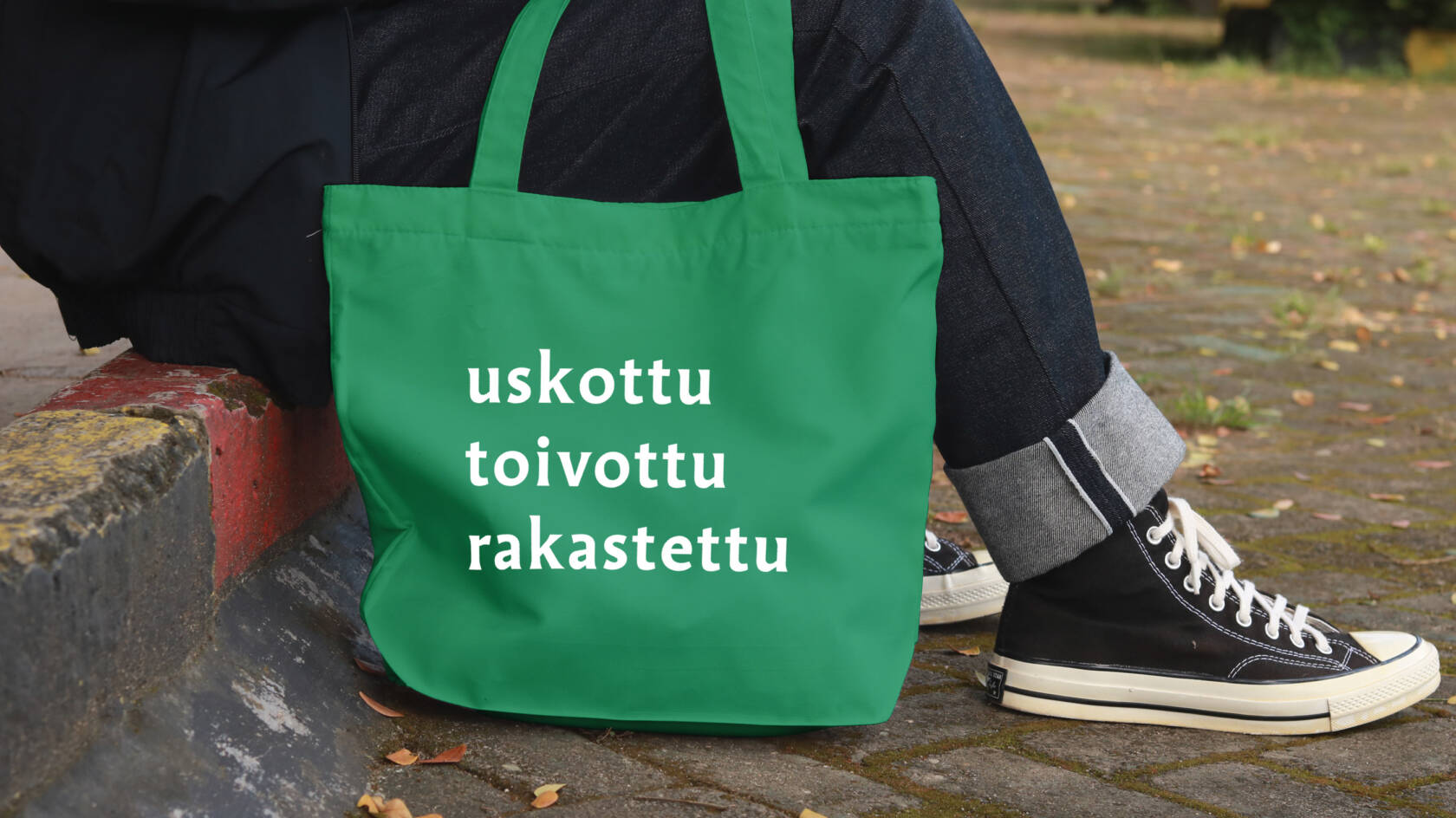 Tienreunalla istuvan ihmisen jalat ja niiden vieressä vihreä kangaskassi, jossa teksti: uskottu toivottu rakastettu.