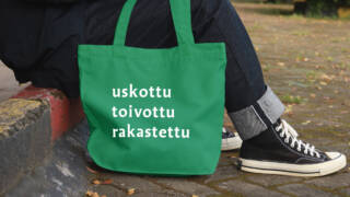 Tienreunalla istuvan ihmisen jalat ja niiden vieressä vihreä kangaskassi, jossa teksti: uskottu toivottu rakastettu.