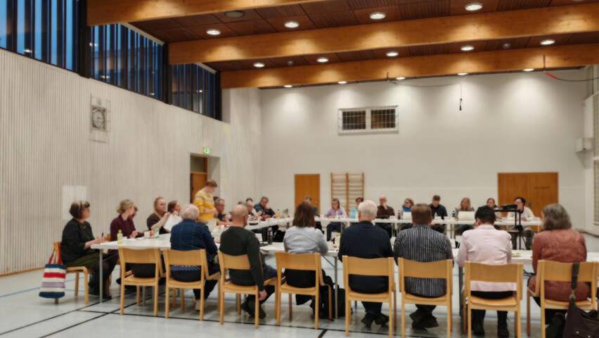 Valaistussa salissa pitkän pöydän ääressä istuu vierekkäin ihmisiä kokoustamassa.